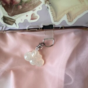 May include: A cloud-shaped charm with a glittery, iridescent finish hangs from a silver-colored clasp. The charm is the focal point against a soft pink background, suggesting a delicate or whimsical theme. The background includes decorative elements with floral and heart motifs.