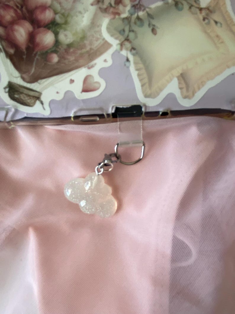 May include: A cloud-shaped charm with a glittery, iridescent finish hangs from a silver-colored clasp. The charm is the focal point against a soft pink background, suggesting a delicate or whimsical theme. The background includes decorative elements with floral and heart motifs.
