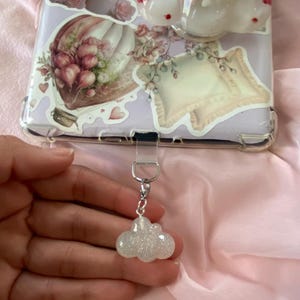 May include: A sparkly, cloud-shaped charm with a silver-colored chain and clasp. The charm is attached to a phone case decorated with floral stickers and a hot air balloon design. The background is a pink fabric.