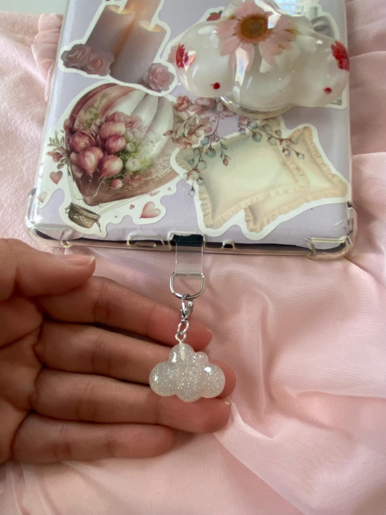 May include: A sparkly, cloud-shaped charm with a silver-colored chain and clasp. The charm is attached to a phone case decorated with floral stickers and a hot air balloon design. The background is a pink fabric.