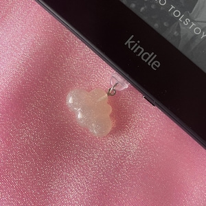 May include: A clear, glittery cloud-shaped charm hanging from a Kindle e-reader. The e-reader is black, with the word "kindle" printed on it. The background is a pink, textured fabric.