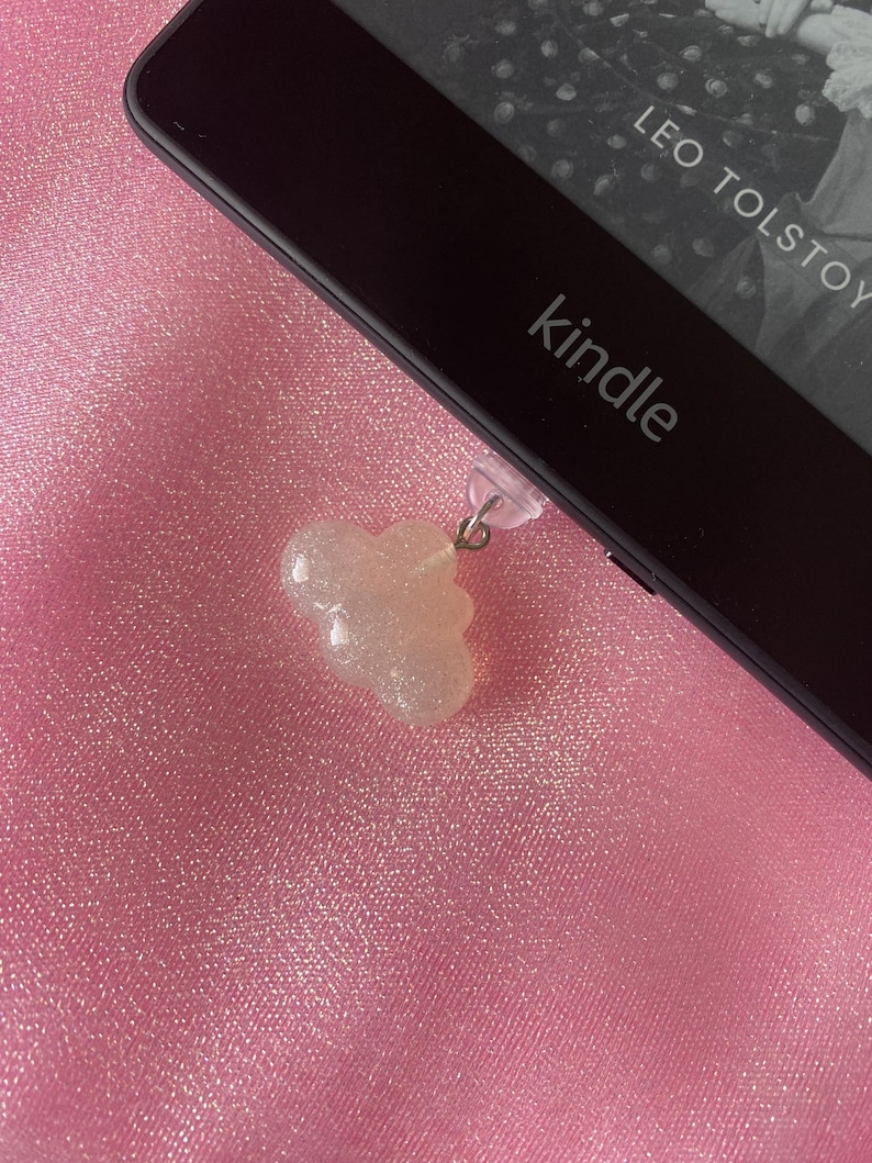 May include: A clear, glittery cloud-shaped charm hanging from a Kindle e-reader. The e-reader is black, with the word "kindle" printed on it. The background is a pink, textured fabric.