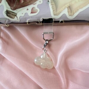 May include: A silver-toned charm with a glittery cloud pendant. The cloud is white and sparkly, hanging from a silver clasp. The background is a soft pink fabric.
