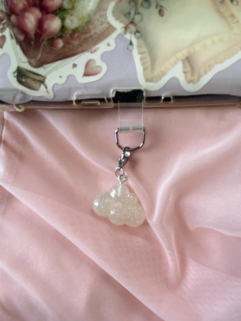 May include: A silver-toned charm with a glittery cloud pendant. The cloud is white and sparkly, hanging from a silver clasp. The background is a soft pink fabric.