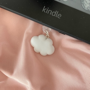 May include: A white cloud-shaped bookmark attached to a black Kindle e-reader. The bookmark is hanging from the top of the Kindle.