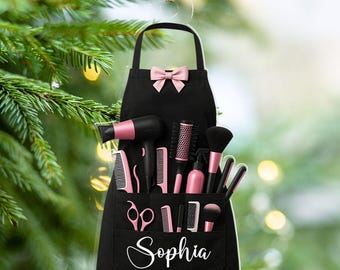 May include: A black apron-shaped Christmas ornament with a pink bow and a pocket filled with pink and black hair styling tools. The name "Sophia" is written in white script on the pocket. The ornament is hanging in front of a Christmas tree.
