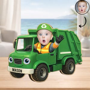 May include: A green toy garbage truck with a baby's face in the driver's seat. The truck has large blue eyes and the name "MASON" on the front. The baby is wearing a green helmet with a recycling symbol and waving.