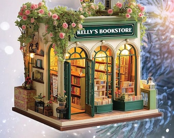 May include: A miniature bookstore model with the text "KELLY'S BOOKSTORE" on the sign. The model features open doors, bookshelves, and pink rose vines. The building has a green and white color scheme, with warm interior lighting.