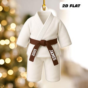 May include: A white karate uniform ornament with a brown belt. The belt is tied in a knot and has the name "JAMES" and the number "2025" printed on it. The text "2D FLAT" is printed in black on the top right corner.