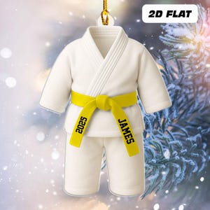 May include: A white karate uniform ornament with a yellow belt. The belt is personalized with the name "JAMES" and the year "2025". The ornament is labeled "2D FLAT".