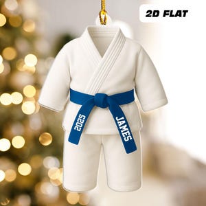 May include: A white karate uniform ornament with a blue belt. The belt is tied in a knot and has the name "JAMES" and the year "2025" printed on it. The ornament is hanging from a gold string and has the text "2D FLAT" in the upper right corner.