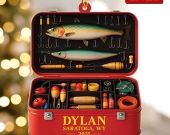 May include: A red, 2D flat Christmas ornament shaped like a fishing tackle box. The top section features two fish and hooks. The bottom section contains various fishing lures and gear. The text "DYLAN SARATOGA, WY 2025" is printed on the front.