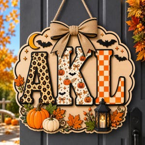 May include: A wooden door sign featuring the letters AKL in leopard print, pumpkin pattern, and orange checkerboard. It is decorated with a burlap bow, a large orange pumpkin, a small white pumpkin, autumn leaves, and a black lantern.
