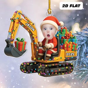 May include: A festive Christmas ornament featuring a baby in a Santa suit operating a yellow excavator adorned with colorful lights. The excavator has the name "JASON" and the year "2025" on it, with a pile of wrapped gifts in the back.