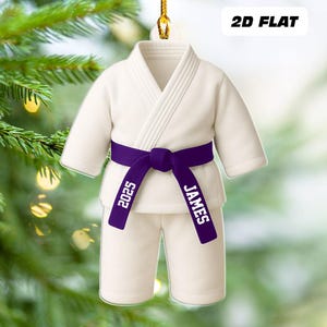 May include: A white karate uniform ornament with a purple belt. The belt is personalized with the name "JAMES" and the year "2025". The ornament is hanging from a gold string, with the text "2D FLAT" in the upper right corner.