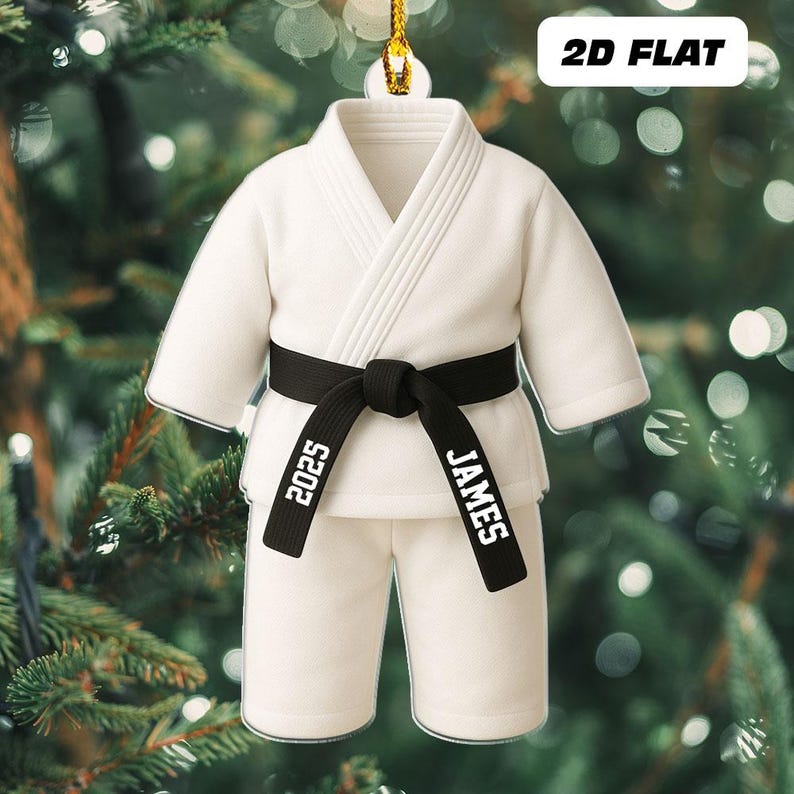 May include: A white karate uniform ornament with a black belt. The belt is tied in a knot and has the name "JAMES" and the year "2025" printed on it. The ornament is hanging from a gold string against a blurred background.