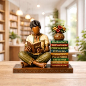 Puede incluir: Una escultura de madera de una persona leyendo un libro con la etiqueta "AMARI", sentada sobre una base de madera. Junto a la figura hay una pila de libros con citas inspiradoras y un jarr&oacute;n con flores. La figura lleva una camisa y pantalones estampados.