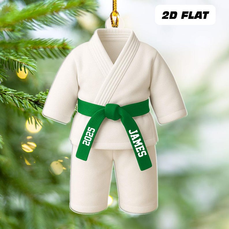 May include: A white karate uniform ornament with a green belt. The belt is personalized with the name "JAMES" and the year "2025". The ornament is hanging from a gold string against a blurred background of a Christmas tree.