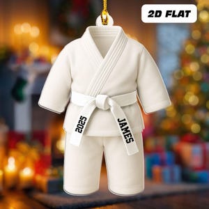 May include: A white karate uniform ornament with a tied belt. The belt has the name "JAMES" and the year "2025" printed on it. The ornament is labeled "2D FLAT".