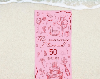 May include: Pink beach towel with a red outline and illustrations of summer-themed items. The text "The summer I turned 50" is displayed, along with "EST. 1975". Includes images of a cake, balloons, and a flamingo float.