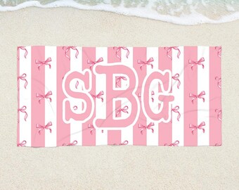 May include: Pink and white striped beach towel with pink bows and the initials "SBG" in a large, stylized font. The towel is rectangular and lies on a sandy beach with the ocean in the background.