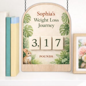 May include: A wooden sign with the text "Sophia's Weight Loss Journey" and the number "3,17" in white tiles, with the word "POUNDS" below. The sign features a watercolor illustration of a rabbit and tropical leaves. It hangs from a chain.