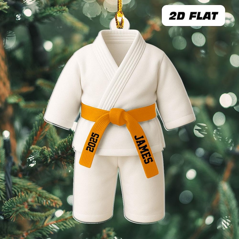 May include: A white karate uniform ornament with an orange belt. The belt is personalized with the name "JAMES" and the year "2025". The ornament is hanging from a gold string, set against a blurred background of Christmas tree branches.