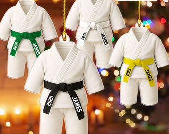 May include: White karate uniform ornaments with black, green, and yellow belts. Each uniform has a personalized belt with the name "JAMES" and the year "2025". The ornaments are hanging with gold strings.