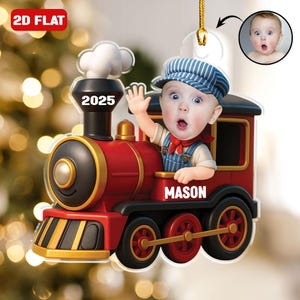 May include: A 2D flat Christmas ornament shaped like a red and gold train with a baby's face in the conductor's seat. The baby wears a blue and white striped cap, overalls, and a red neckerchief. The ornament has the text "2025" and "MASON".
