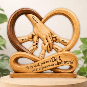 May include: A wooden heart-shaped sculpture with a dark brown and light brown finish. The sculpture features a hand holding smaller hands, with names etched on each small hand. The base has the text: "To the world you are a Dad, but to us you are our whole world."