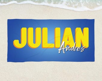 May include: A blue beach towel with the name "JULIAN" in large yellow letters with an orange shadow. The name "Andres" is written in white script below "JULIAN". The towel is on a sandy beach with ocean waves in the background.
