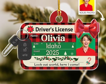May include: A personalized Christmas ornament shaped like a driver's license, featuring the name "Olivia" and "Idaho" with a 2025 issue date. The ornament includes a car key and a photo, with the text "Look out world, here I come!" and a red bow.