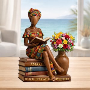 May include: A decorative figurine of a woman reading, seated on a stack of books. The figurine wears a colorful dress and head wrap. The books read "AMARI" and "BLACK. EDUCATED. POWERFUL." A vase of flowers sits beside her.