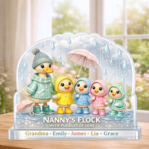 May include: A decorative acrylic display featuring a duck and five ducklings in raincoats and boots under a rainy sky. The text reads "Nanny's Flock with Puddles of Love" with names below. The ducklings wear yellow, blue, pink, and green raincoats.
