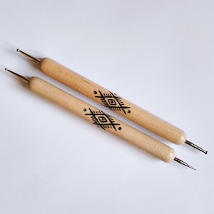 May include: Two wooden dotting tools with metal tips. The light-colored wood handles feature a black geometric design. These tools are used for creating dots and patterns in various craft projects.