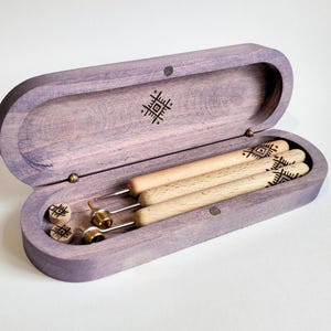 May include: A wooden box with a purple stain, containing four wooden tools with a geometric pattern. The box has a hinged lid and a small latch.