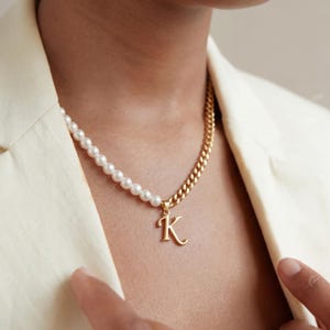 Puede incluir: Un collar dorado con una hilera de perlas a la izquierda y una cadena de eslabones a la derecha. Un colgante dorado con la letra "K" cuelga de la cadena. El collar se lleva sobre una blazer color crema.