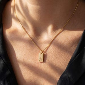 May include: A gold-colored necklace with a rectangular pendant. The pendant features the letter "A" engraved on it. The necklace is a delicate chain, and the pendant hangs at the center. The necklace is shown against a dark background.