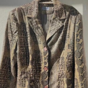 May include: A long snakeskin-effect blazer in brown and tan hues. The jacket features a notched collar, long sleeves, and a three-button fastening. It is displayed on a wooden hanger against a plain backdrop.