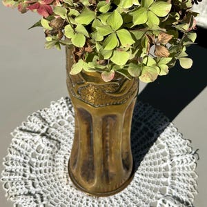 May include: A brass vase with an ornate design, featuring a bouquet of green and pink hydrangeas. The vase is sitting on a white crocheted doily.