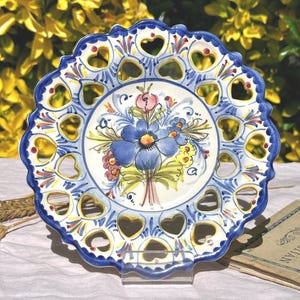 Puede incluir: Plato decorativo de cerámica con borde festoneado y recortes en forma de corazón. El plato presenta un diseño floral pintado a mano en azul, amarillo y rosa. El borde está adornado con un borde azul. El plato se exhibe sobre un soporte transparente.