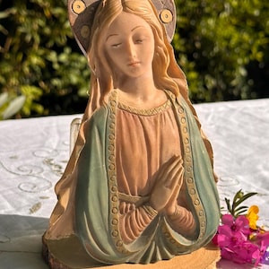 Puede incluir: Una figurita vintage de la Virgen María con un halo, vestida con una túnica azul y verde, con las manos juntas en oración. La figurita está sobre una base de madera.
