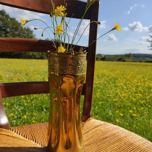 May include: A brass vase with a decorative rim holds yellow wildflowers. The vase sits on a woven chair, with a field of yellow flowers and a blue sky in the background. The vase is approximately 25 cm tall.