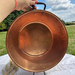 Peut inclure: Une poêle ronde de couleur cuivre avec une poignée noire et un support assorti. La poêle a un design large et peu profond et est faite d'un métal brillant. La poêle est soutenue par un support en métal noir. Le fond est un champ avec un ciel bleu.