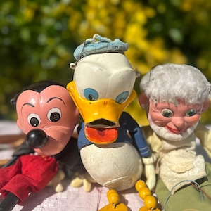 Puede incluir: Tres marionetas de personajes vintage: Mickey Mouse con pantalones cortos rojos, el Pato Donald con un traje de marinero azul y un anciano con pelo blanco. Las marionetas tienen caras pintadas y están juntas.