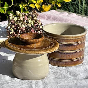 Può includere: Un burriera in ceramica con coperchio e contenitore separato. Il coperchio è marrone scuro con un motivo circolare. La base è beige chiaro. Il contenitore è marrone più scuro con strisce orizzontali.