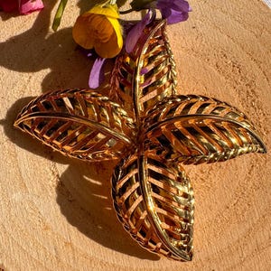 Puede incluir: Un broche de metal dorado en forma de flor estilizada con cuatro hojas. Las hojas están hechas de finas tiras de metal entrelazadas.