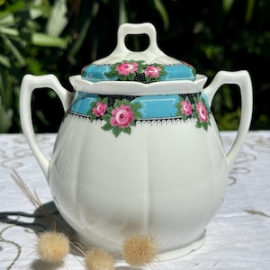 Peut inclure: Sucrier en céramique blanche avec couvercle, orné d'une bande turquoise ornée de roses roses et de feuilles vertes. Le bol a deux poignées et un design côtelé. Le couvercle a une poignée et un motif floral. Le sucrier est sur une surface blanche.