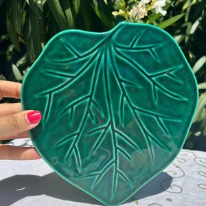 May include: A glossy, emerald green ceramic leaf-shaped dish. The dish features detailed veining and a smooth, curved surface. The leaf design is a decorative accent for serving or display.