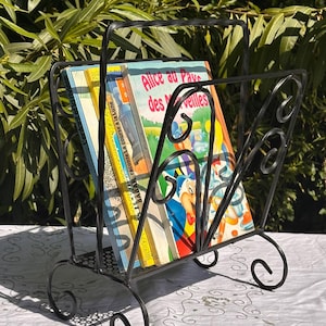 Può includere: Porta riviste in metallo nero con un design a volute decorative. Il porta riviste contiene una pila di libri, tra cui uno intitolato "Alice nel Paese delle Meraviglie".